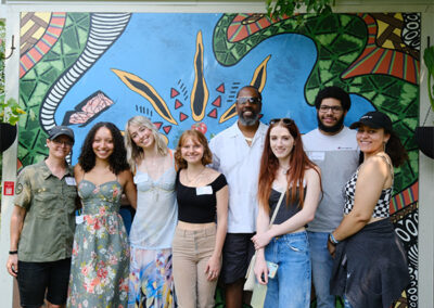A group of six students stands outside in front of a mural with two instructors.
