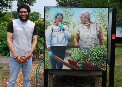 Isaic Pulliam stands in a garden next to a realistic painting of a child and their grandmother with a wheelbarrow of vegetables.