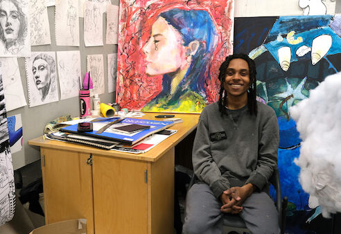 Brandon Luster sits in his studio, surrounded by his pencil and pastel drawings. On the desk next to him is a large pile of art suppplies.