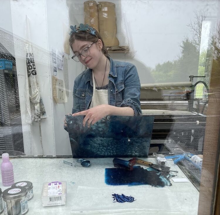 Artist Julia Flock creating work in the print studio. She's holding a see-through panel with ink on it, and some rich blue ink is rolled out on a surface in front of her.