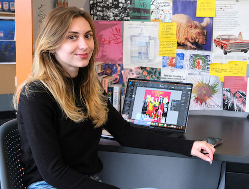 Lauren Blood sits in her studio space, turned away from her desk to face the camera. Her laptop is open with the screen displaying her digital workspace, and a collage of personal work and inspirational materials are on the wall behind her.