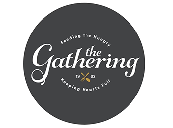 Grey circle logo for The Gathering with the organization name in white decorative font through the middle. The tagline curves above and below the middle of the name, reading "Feeding the Hungry / Keeping Hearts Full." Between the organization name and the bottom half of the tagline is the established year "1982" with a gold illustration of a crossed fork and spoon between the 19 and 82.